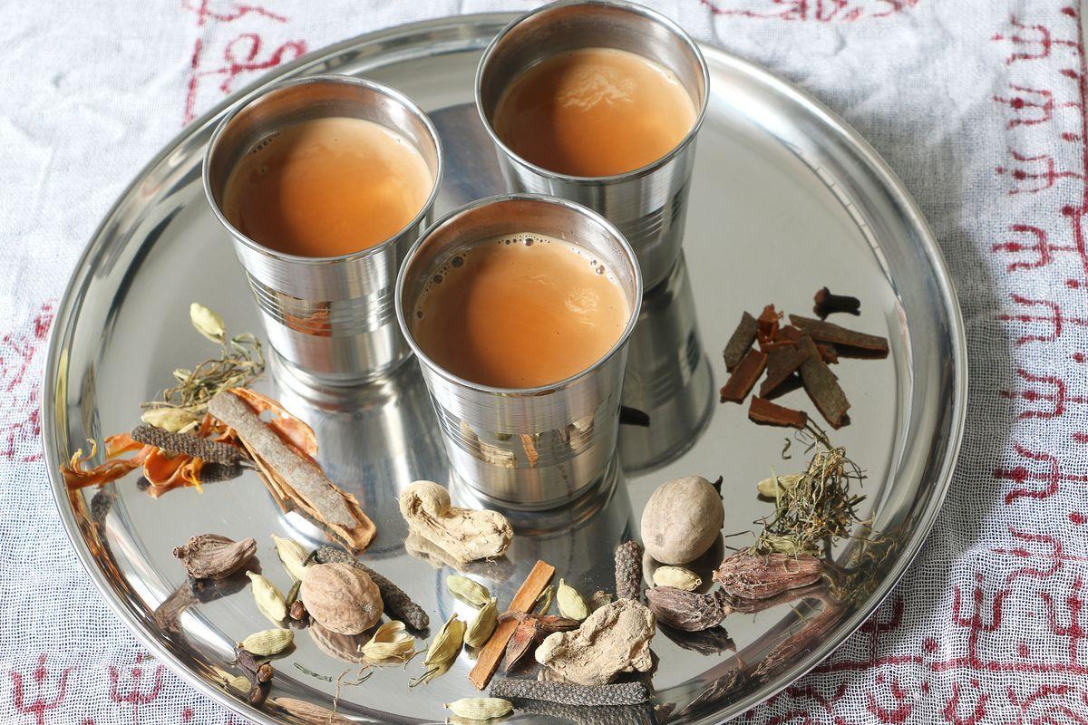 Three stainless steel cups of chai on a metal tray with various aromatic spices scattered around.