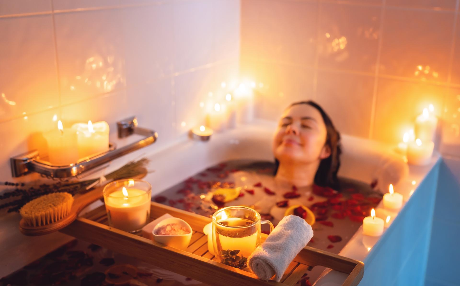 Young woman enjoying spiritual aura cleansing rose flower bath with rose petals and candles during full moon ritual. Body care and mental health routine.