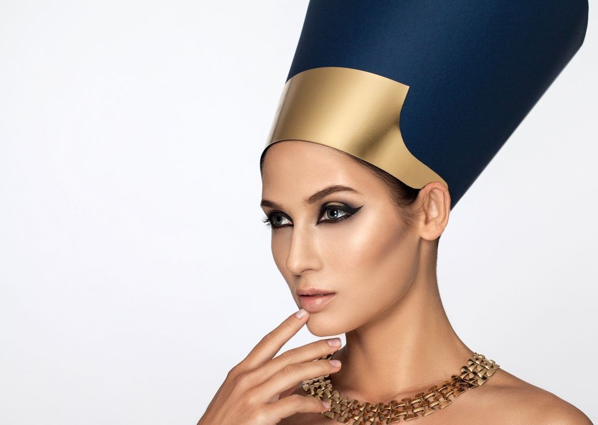 Woman wearing an Egyptian-inspired headdress and necklace, with dramatic makeup and a thoughtful expression.